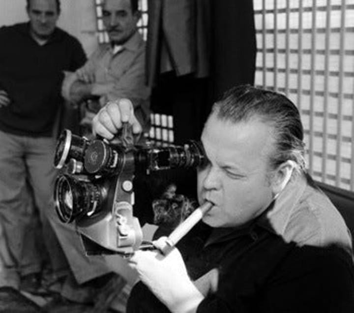 Photo of Orson Welles smoking while he shoots a shot on location.